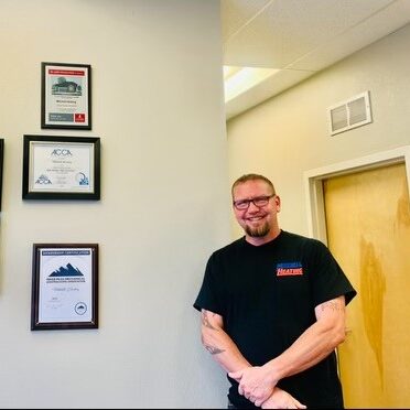 Portrait of Sam Moore, General Manager at Mitchell Heating, wearing glasses and standing next to a wall with framed certificates